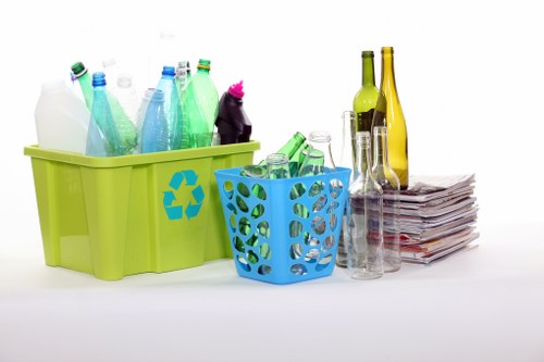 The image displays a collection of empty glass bottles and plastic containers, arranged on a white background. On the left, a large, lime-green plastic bin featuring a recycling symbol is filled with transparent plastic bottles in shades of blue, green, and clear, some with caps and labels. Next to it, a smaller, blue plastic basket with circular cutouts holds more plastic bottles, mostly green, with some lying horizontally. To the right, an assortment of glass bottles in various shapes and sizes, including tall, slender green and amber-colored bottles, are stacked or leaning against each other, with some placed on a neat pile of crumpled newspapers or magazines. The scene appears to be set indoors, possibly in a recycling or waste collection area, illustrating typical materials collected for rubbish removal services in Brixton, reflecting environmentally conscious disposal practices common in the London area. Business Waste Removal Brixton provides comprehensive rubbish management, as suggested by the visual emphasis on recyclable containers and bottles in this arrangement.