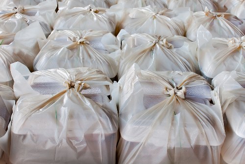A closely packed arrangement of clear plastic rubbish bags filled with waste material, tied securely at the tops with twist ties, resting on a flat surface in an outdoor setting. The bags appear to contain a mix of household or commercial waste, with some showing slight bulges or uneven shapes due to the contents inside. The surface beneath the bags is not visible, but the bags are arranged in orderly rows, suggesting they are prepared for collection or disposal. The background is blurred and neutral, focusing attention on the bags. This image is representative of waste removal services, such as those provided by Business Waste Removal Brixton, in a typical urban or suburban environment within the Brixton area, highlighting the typical process of waste collection in a professional and organized manner.