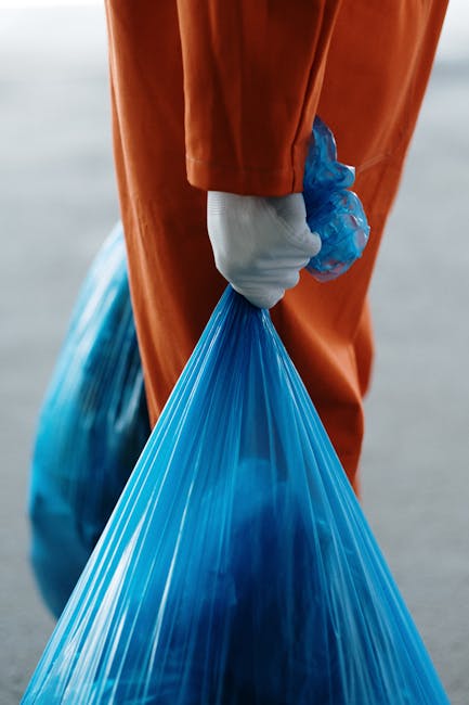 A person dressed in high-visibility orange workwear with reflective strips and an orange cap is seen from above, standing on a paved sidewalk next to a curb. They are holding a broom with a wooden han