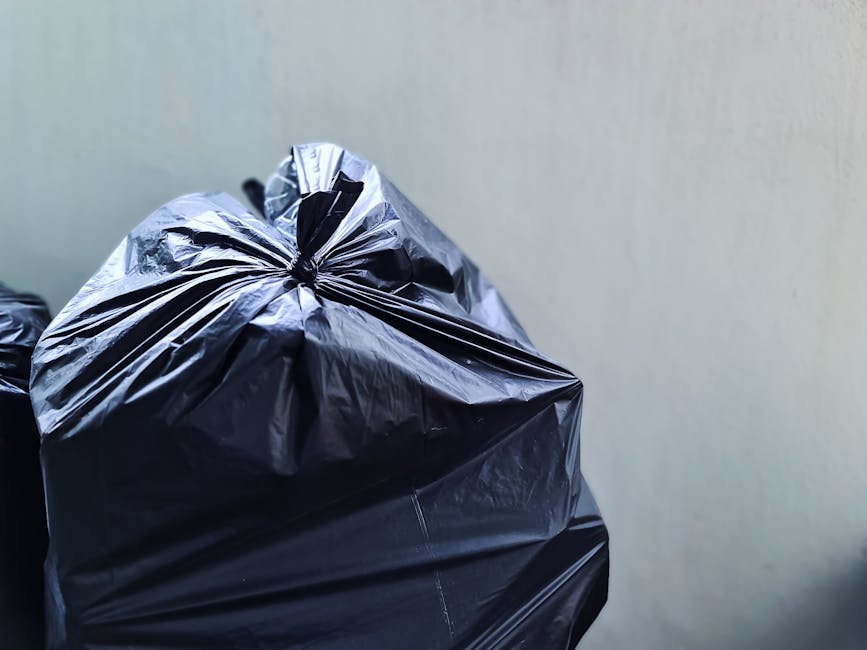 Close-up view of a large black plastic trash bag made from shiny, crinkled polyethylene material, tightly knotted at the top, positioned on the left side of the image. The bag appears full and is rest